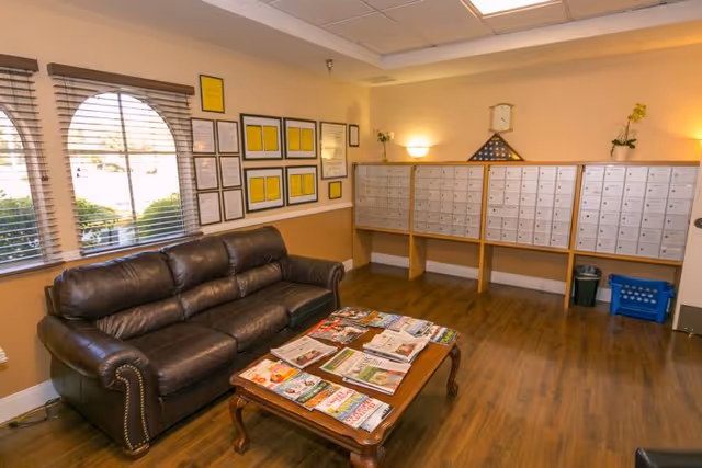 A seating area with a leather sofa and a coffee table covered in magazines next to a wall of mailboxes in a common room.