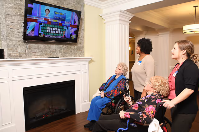Two elderly women in wheelchairs and two female caregivers are watching a television mounted above a fireplace in a cozy room. The TV screen shows a game with the text 'SOLO ROUND' and 'Turns Left'. The room has wooden flooring, white pillars, and light-colored walls.