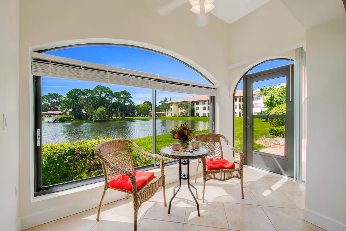 Sunlit sitting area with two wicker chairs and a small table by large arched windows overlooking a lake and neighboring buildings.