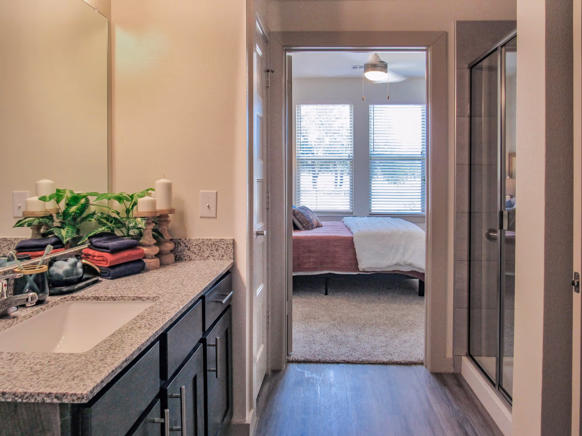 View of a bathroom with a granite countertop, sink, and decorative candles and plants on the left. A glass-enclosed shower is on the right. The bathroom opens into a bedroom with a bed, pillows, and two large windows with blinds letting in natural light.