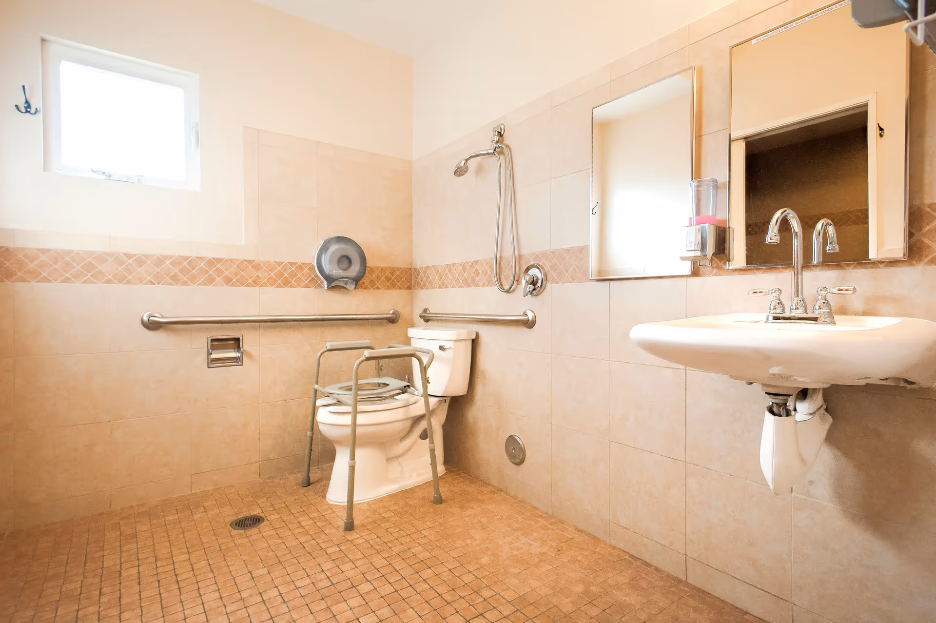Accessible tiled bathroom with a toilet fitted with a safety frame, wall grab bars, shower head, and a wall-mounted sink.