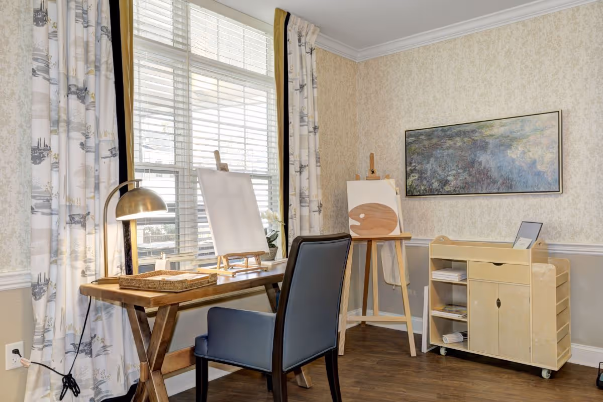 A bright and cozy art corner in a senior living facility featuring a wooden desk with a lamp, a chair, two easels with blank canvases, a rolling storage cart with art supplies, and a large window with patterned curtains allowing natural light to fill the room.