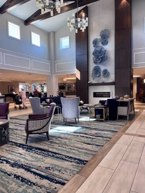 Spacious and well-lit assisted living facility common area with high ceilings, large windows, and exposed wooden beams. The room features a modern fireplace with artistic wall decor above it, several upholstered chairs and sofas arranged around coffee tables on a patterned rug. In the background, a person is walking with a walker, and there is a piano and other seating areas visible.