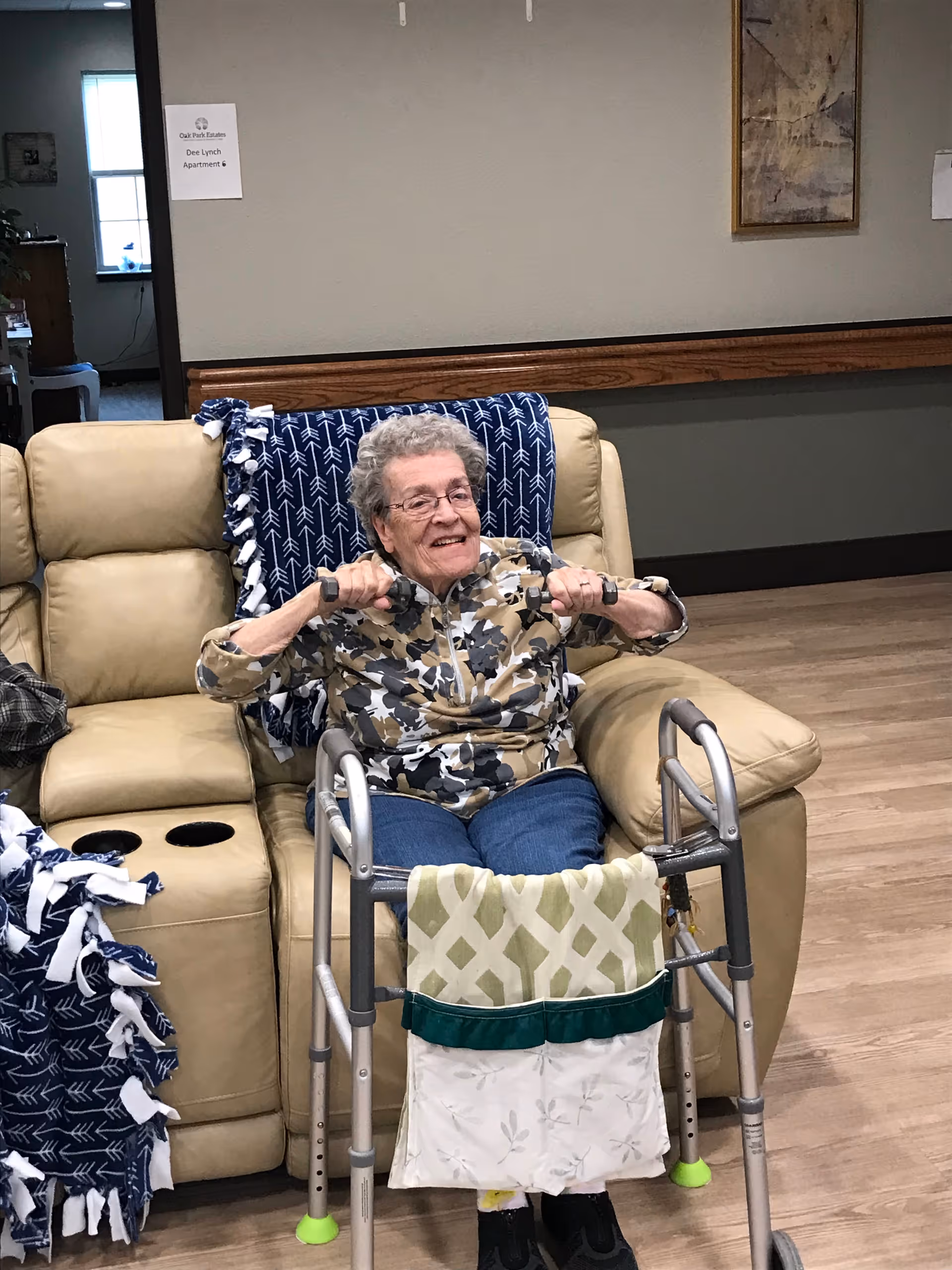 An elderly woman sitting on a beige leather couch in a living room area, smiling and holding small hand weights. She has a walker in front of her with a fabric pouch attached. The room has wooden flooring, a blue patterned blanket on the couch, and a sign on the wall that reads 'Oak Park Estates Dee Lynch Apartment 6'.