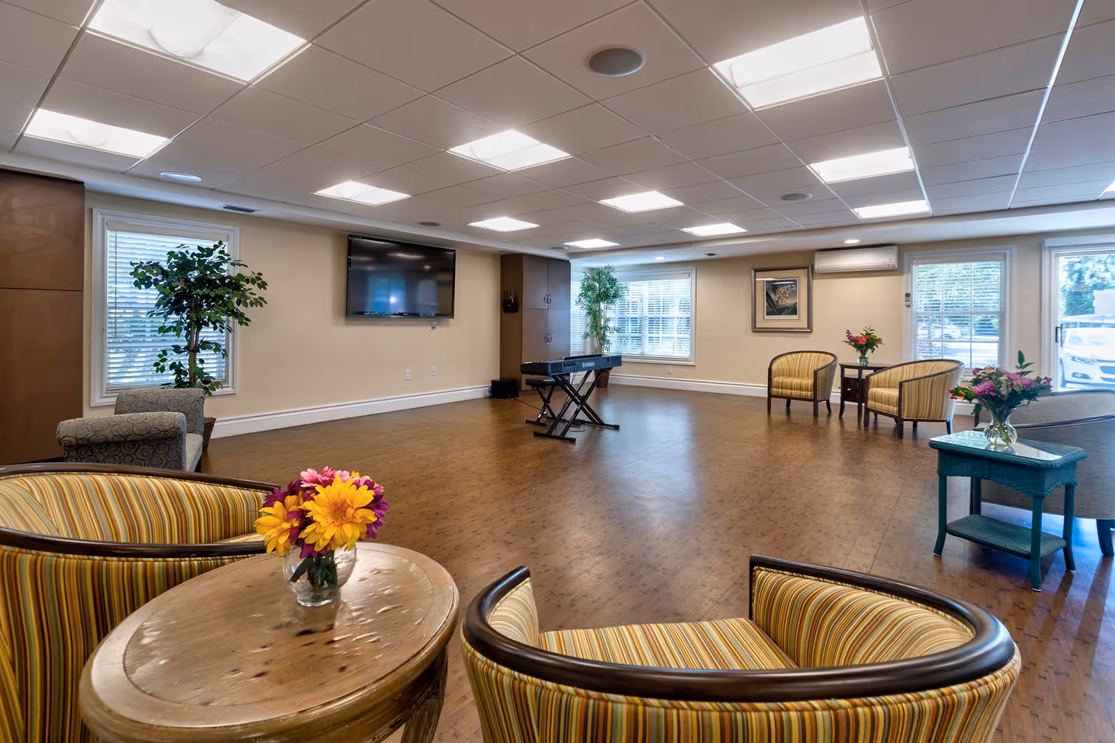 A spacious, well-lit common area with wooden flooring, several striped armchairs arranged around small tables with flower vases, a keyboard on a stand, a wall-mounted flat screen TV, large windows with blinds, and potted plants near the windows.
