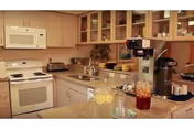 A kitchen area with beige cabinets, a white microwave, and stove. There is a granite countertop with a glass container of iced tea and a glass of lemon water. Various kitchen appliances and utensils are visible, including a coffee maker and toaster.