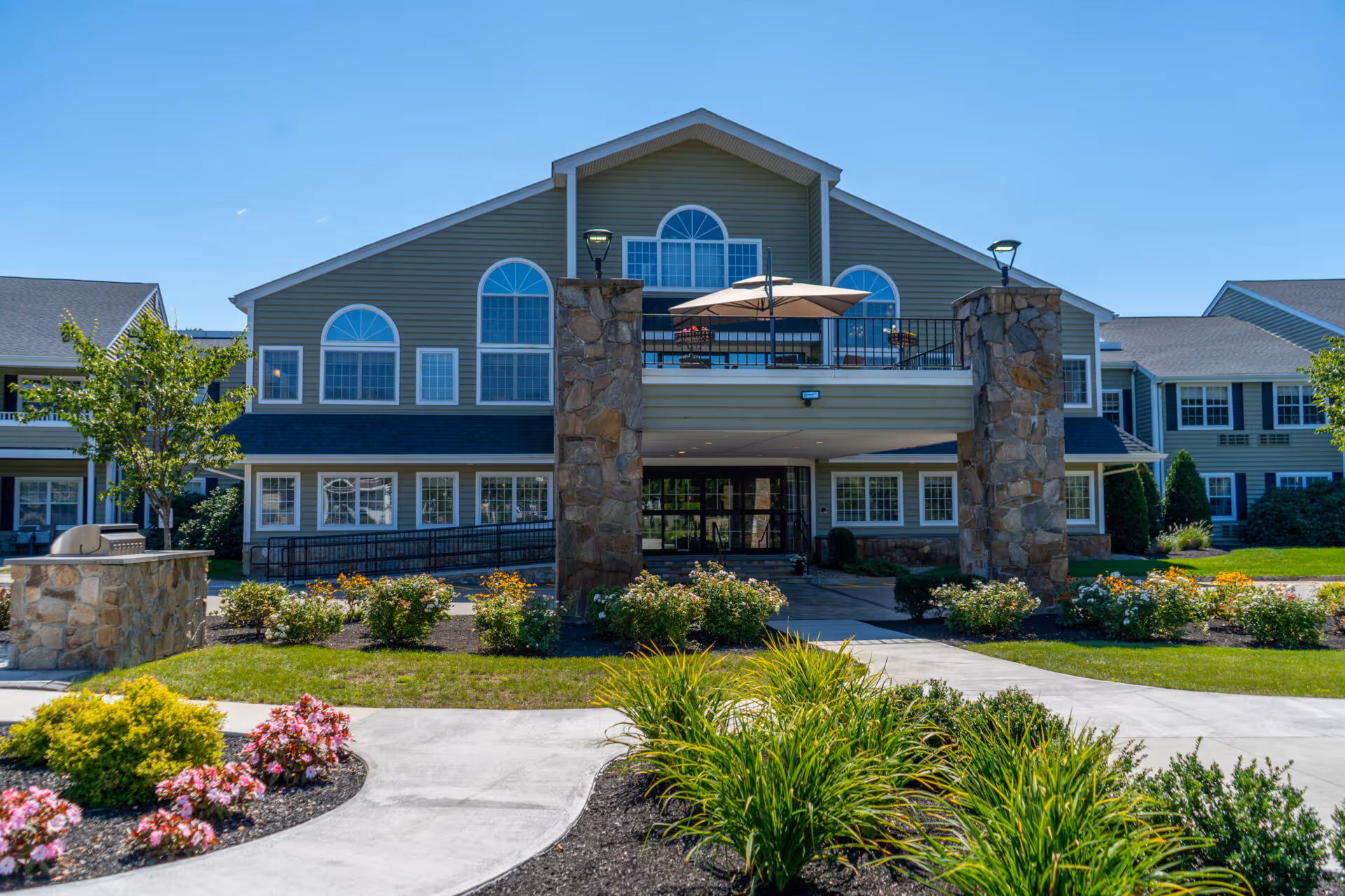 Exterior view of a senior living facility building with stone pillars at the entrance, large windows, a balcony with an umbrella, and well-maintained landscaping including bushes and flowers along a curved concrete walkway.