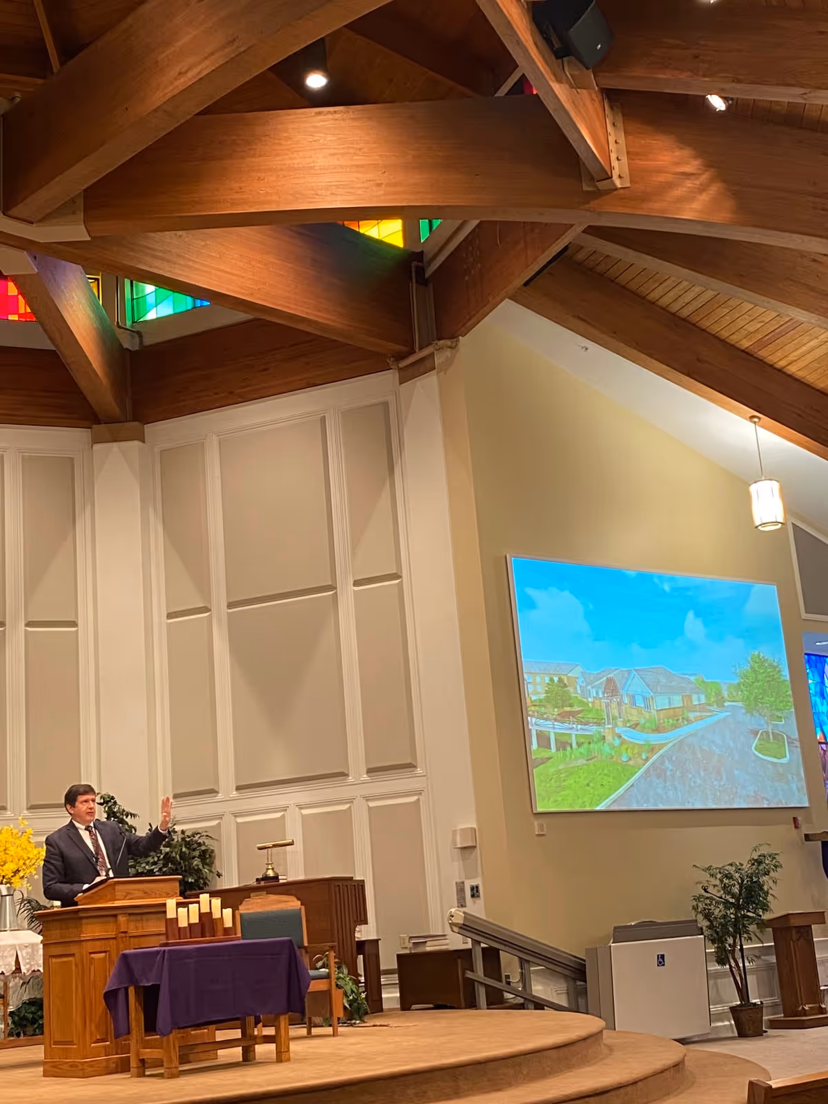 A man in a suit stands at a wooden podium inside a large room with high wooden beams and stained glass windows near the ceiling. Behind him is a wall with white paneling and a large screen displaying an image of a building and surrounding greenery. There are plants and a table covered with a purple cloth on the stage.
