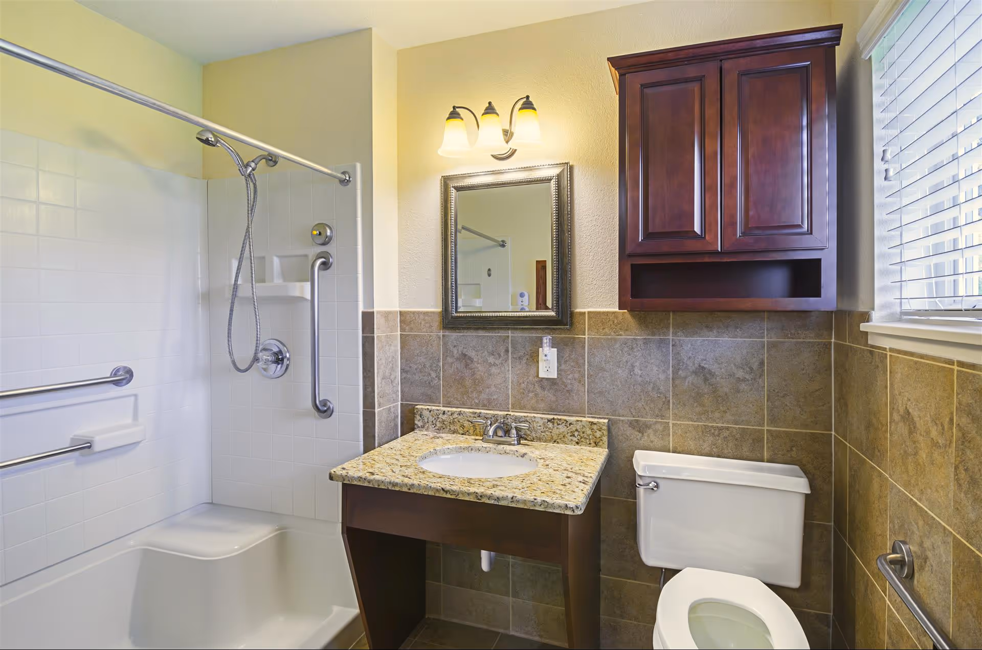 A bathroom with a white bathtub and shower combination featuring grab bars and a handheld showerhead. There is a granite countertop with a sink, a mirror above it, and a wooden cabinet mounted on the wall. The walls are tiled halfway up and painted light yellow above the tiles. A window with blinds is visible on the right side.