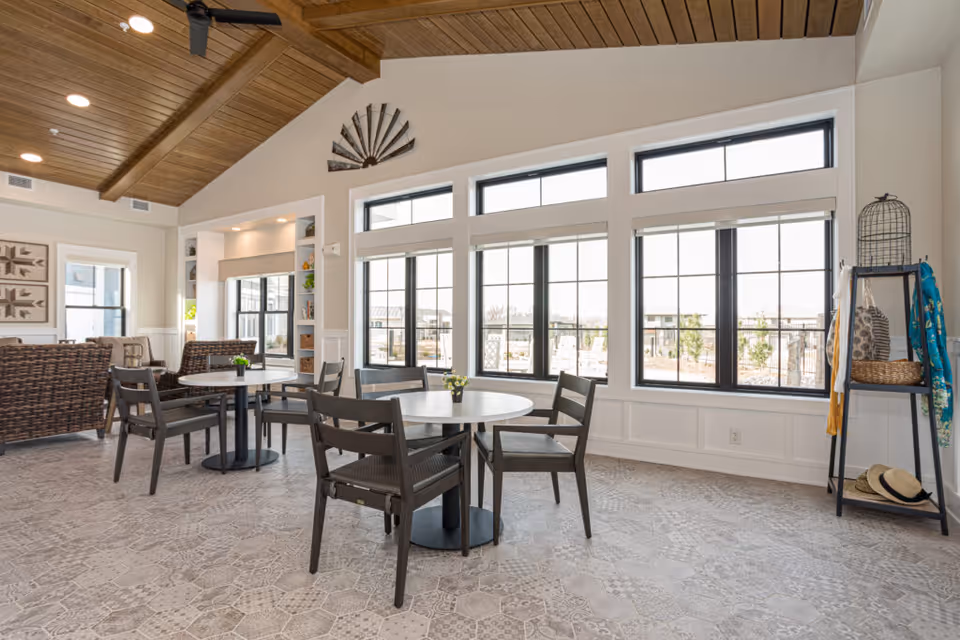 A bright and spacious common area with large windows letting in natural light. The room features round tables with chairs, wicker seating, and decorative items such as a wall fan and a shelf with hats and scarves. The ceiling has wooden beams and a ceiling fan.