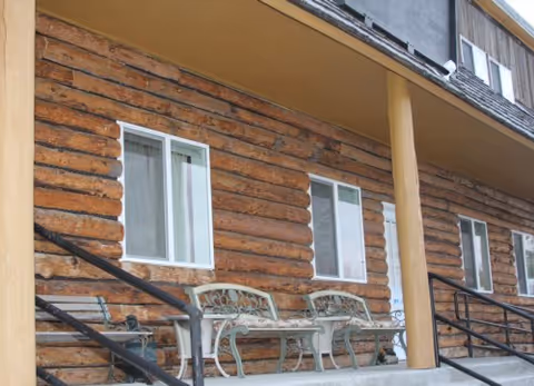 Front exterior porch of a log-sided building with two decorative metal benches, three windows, wooden support posts, and handrails.