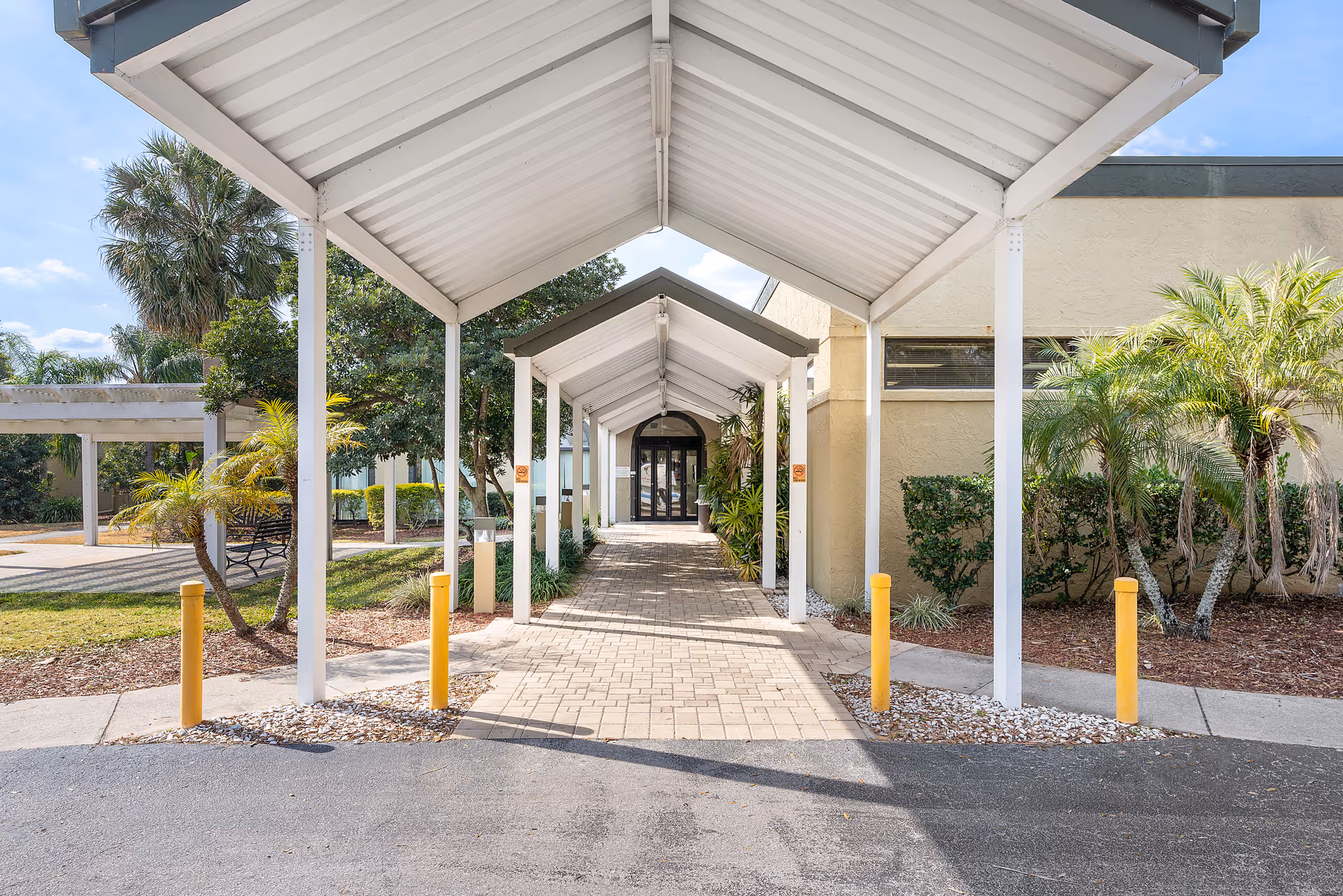 Covered walkway with white metal roof and columns leading to the entrance of a building, surrounded by palm trees and landscaping under a partly cloudy sky.