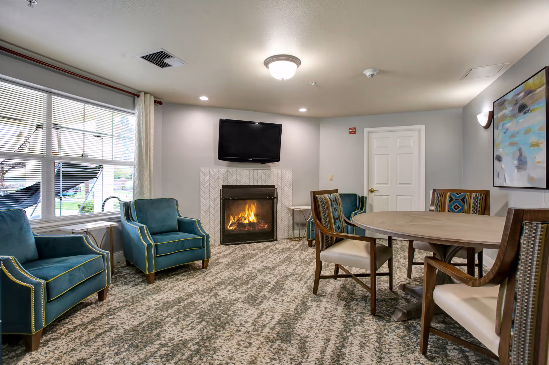 A cozy senior living common area with a lit fireplace beneath a wall-mounted flat screen TV. The room features three teal armchairs with yellow piping, a round wooden table with four upholstered chairs, a large window with blinds and curtains, and a colorful abstract painting on the wall.