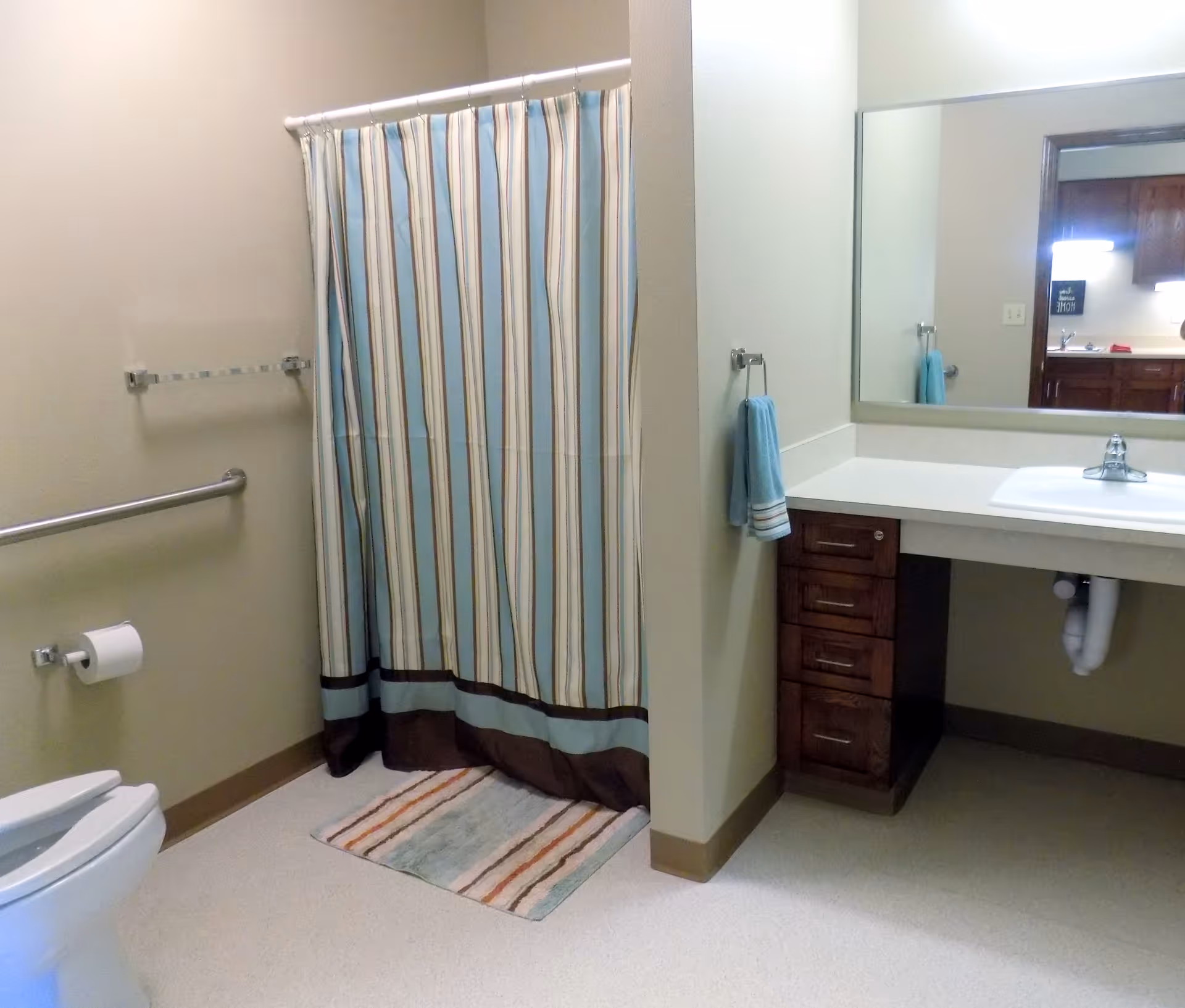 A bathroom with a toilet, a striped shower curtain, a small rug, a sink with a large mirror above it, and a towel hanging on a rack. The bathroom has beige walls and a light-colored floor.