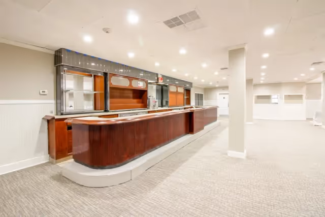 A spacious interior room with a long wooden serving counter and glass display shelves behind it, illuminated by ceiling lights. The room has light-colored walls and carpeted floors, with columns and an open layout.