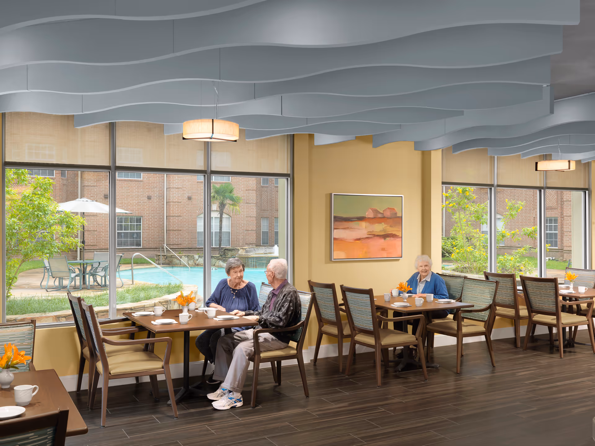 A dining area in a senior living facility with large windows showing an outdoor pool and garden. Two elderly people are sitting at one table engaged in conversation, and another elderly woman is seated at a nearby table. The room has wooden floors, modern chairs, and a wavy ceiling design with pendant lights. A colorful painting hangs on the wall.