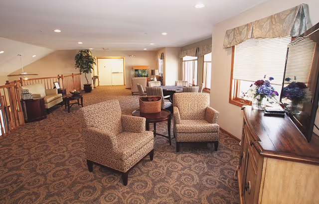 A spacious, well-lit common area with patterned carpet, two upholstered armchairs around a small round table, additional seating and tables in the background, large windows with valances, a wooden cabinet with a flat-screen TV and flower arrangements, and a potted plant near a railing.