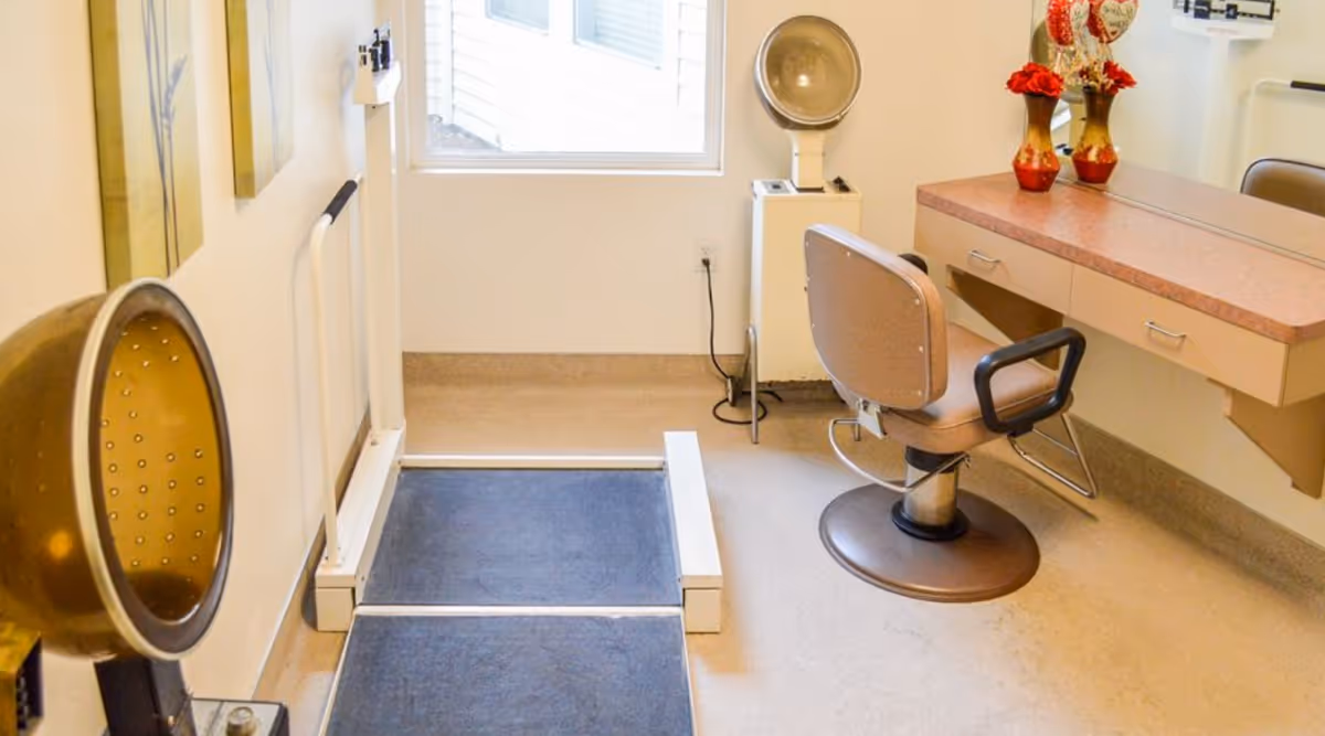 Interior of a salon room in a senior living facility with a salon chair, hair drying hood, a vanity with a mirror and decorative vases with red flowers, and a black mat on the floor near the wall.