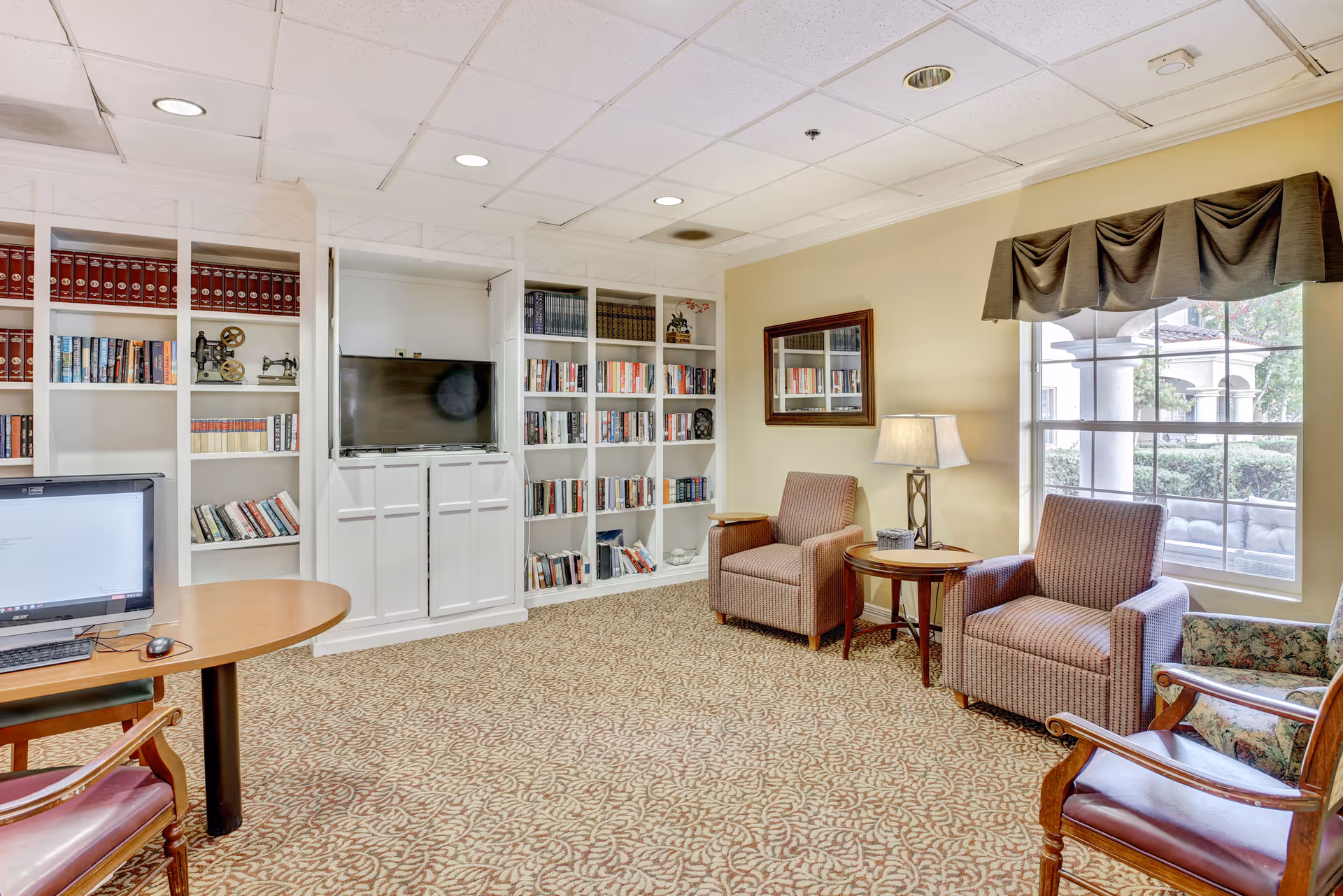 A cozy living room area with patterned carpet, two upholstered armchairs, a small round wooden table with a lamp, a large window with a valance, built-in white bookshelves filled with books and decorative items, a flat-screen TV, and a wooden desk with a computer and chair.