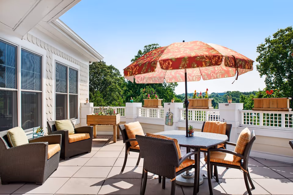 Sunny outdoor patio with a round table, wicker chairs, potted planters and a floral umbrella overlooking trees.