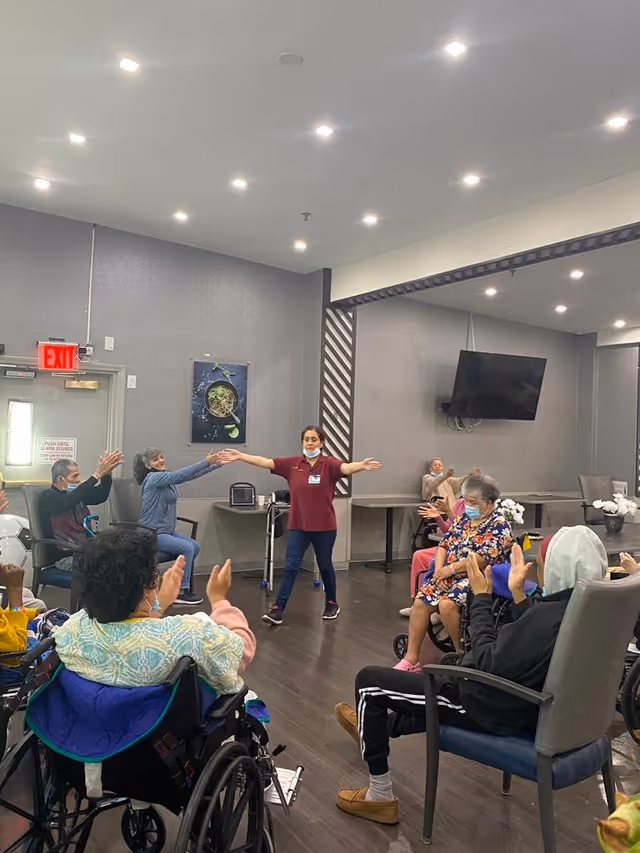 A group of elderly people, some in wheelchairs, participating in a seated exercise or activity led by a staff member in a maroon shirt inside a spacious room with gray walls and wooden flooring. The staff member stands in the center with arms extended, while the elderly participants mimic the movements. The room has a wall-mounted TV, tables, chairs, and a visible exit door with a red exit sign.