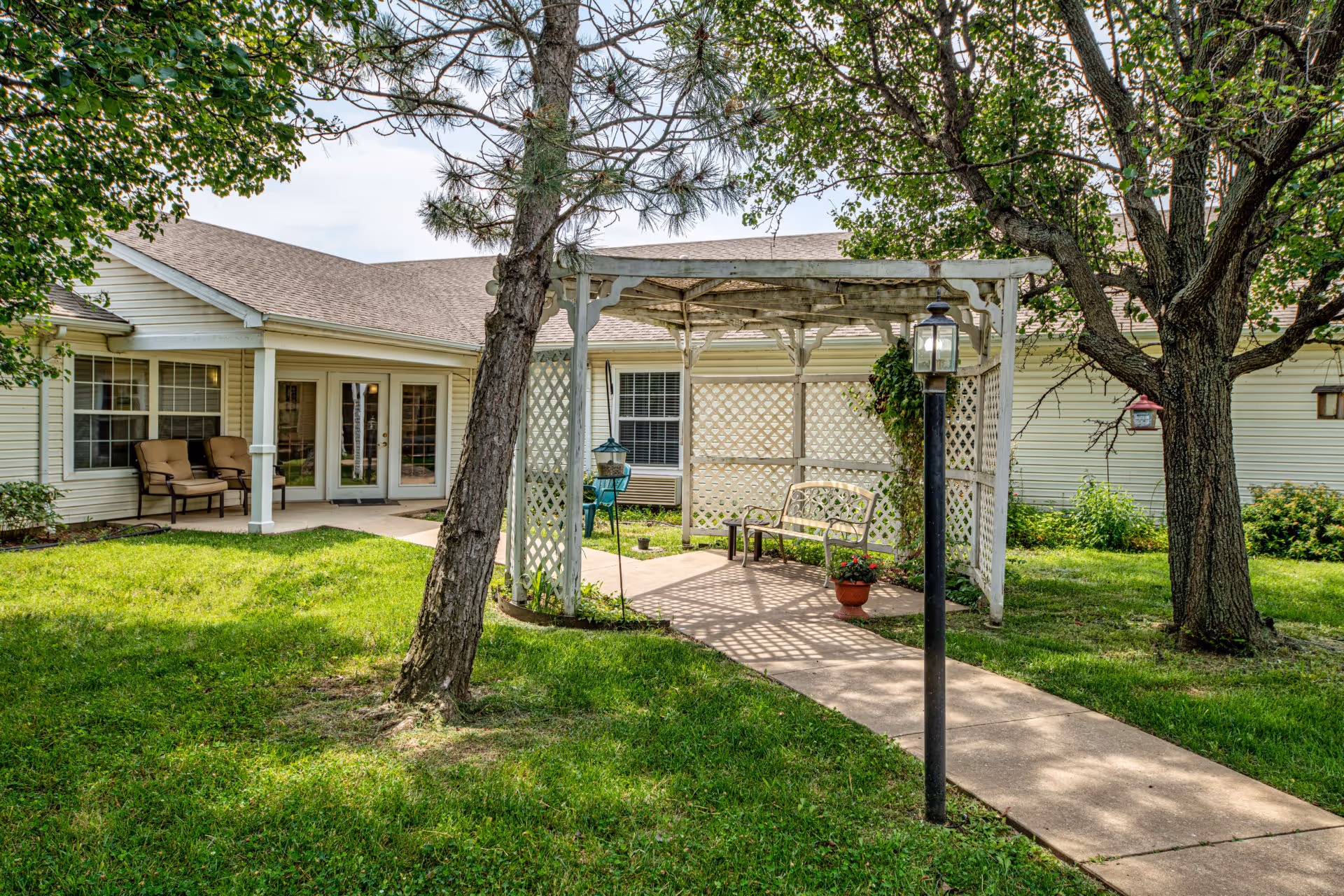 Outdoor garden area at Creekside Village featuring a white pergola with lattice sides, a bench, potted plants, a black lamp post, and trees surrounding a concrete pathway leading to a building with beige siding and glass doors.