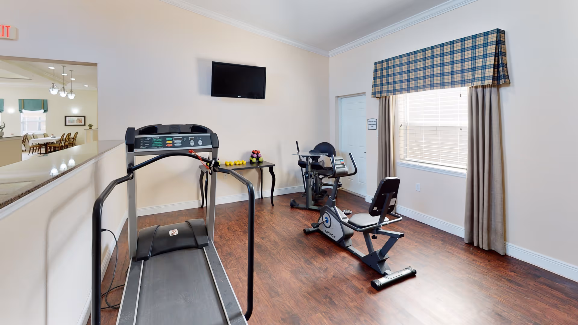 Small indoor fitness room with a treadmill, recumbent bike, wall-mounted TV and a table holding dumbbells.