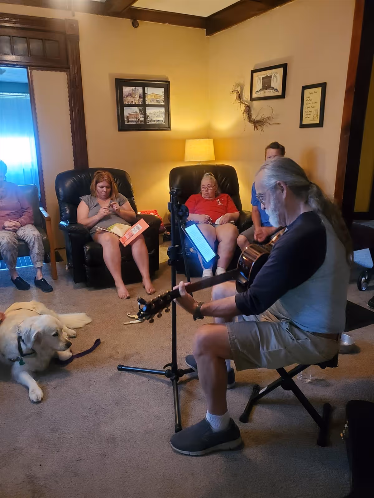 A group of people sitting in a cozy living room with beige walls and carpet. One man with gray hair and a ponytail is playing an acoustic guitar while seated on a small stool. Several people are seated on armchairs and a couch, listening or engaged in quiet activities. A large white dog lies on the carpet near the group. The room is softly lit by a table lamp, and framed pictures hang on the wall.