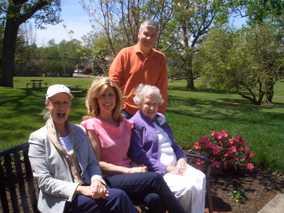 Four people sitting and standing on a bench in a sunny outdoor garden area with green grass, trees, and blooming flowers. Three women are seated on the bench, and one man is standing behind them. They are smiling and enjoying the pleasant weather.
