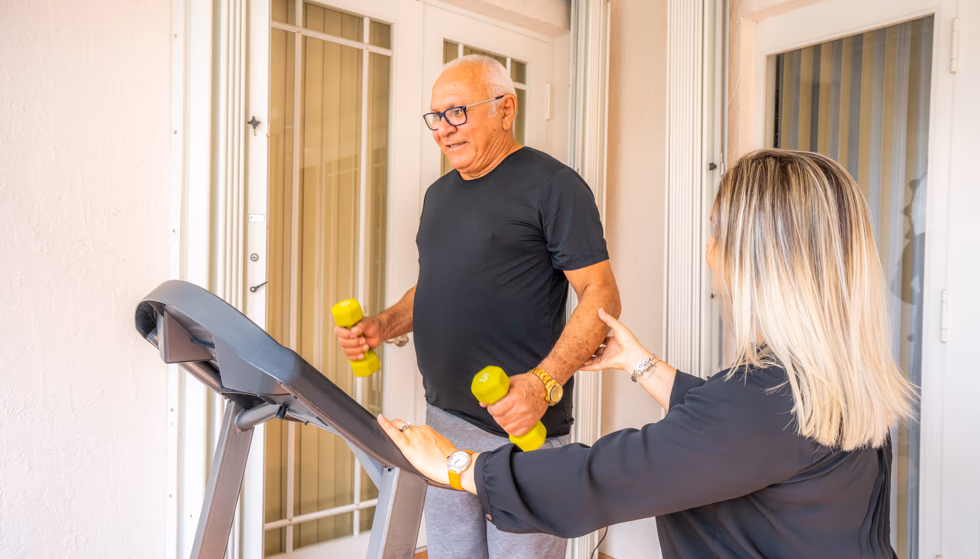 An elderly man wearing a black t-shirt and gray pants is exercising with yellow dumbbells while standing on a treadmill. A woman with blonde hair, wearing a black long-sleeve shirt, is assisting or coaching him. They are indoors near a set of glass doors with white frames.