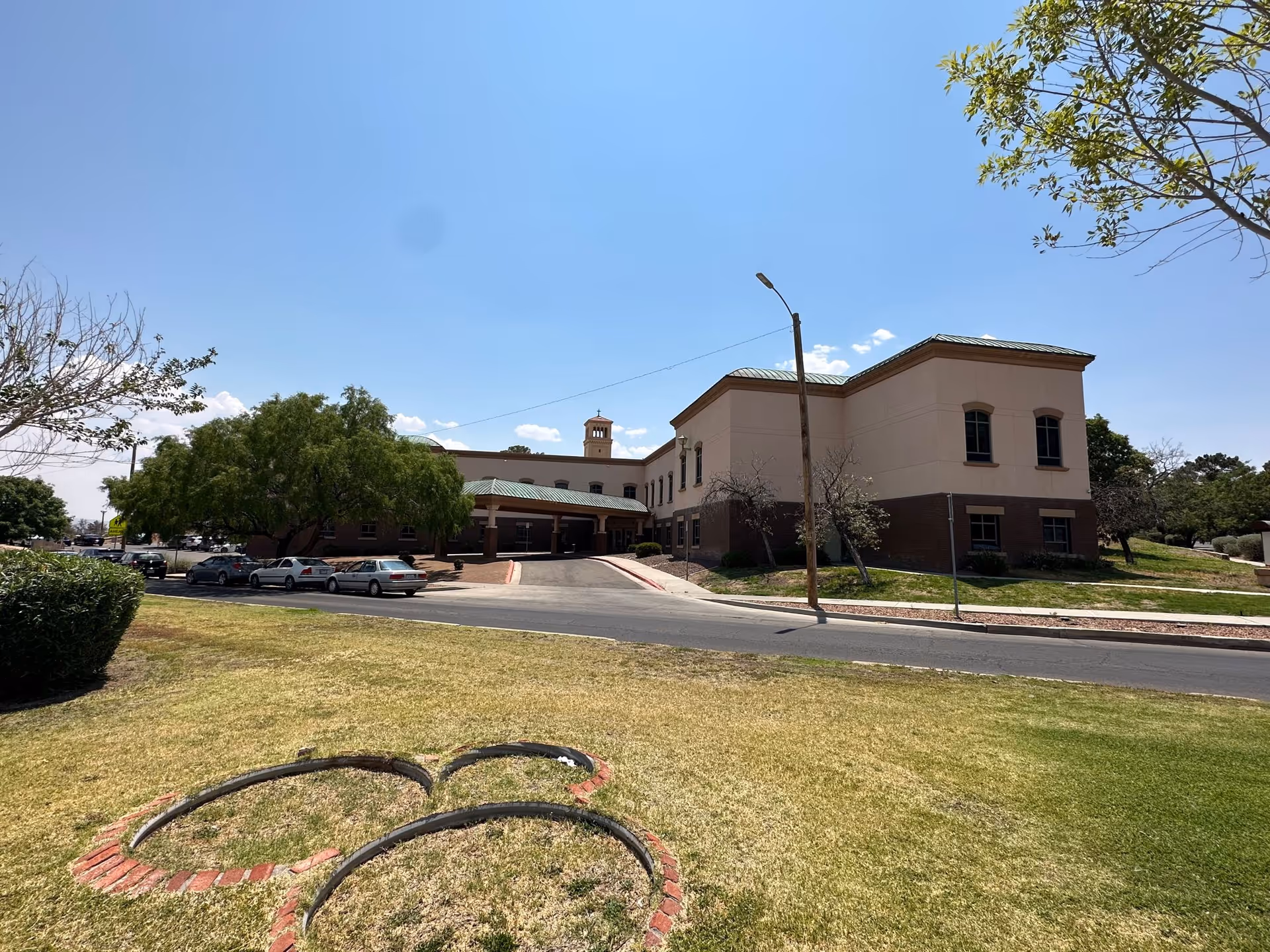 Exterior view of a two-story building with a green roof and beige walls, surrounded by trees and a grassy area with a flower bed shaped like Mickey Mouse's head. Several cars are parked along the driveway leading to the building entrance under a covered drop-off area. The sky is clear and blue.