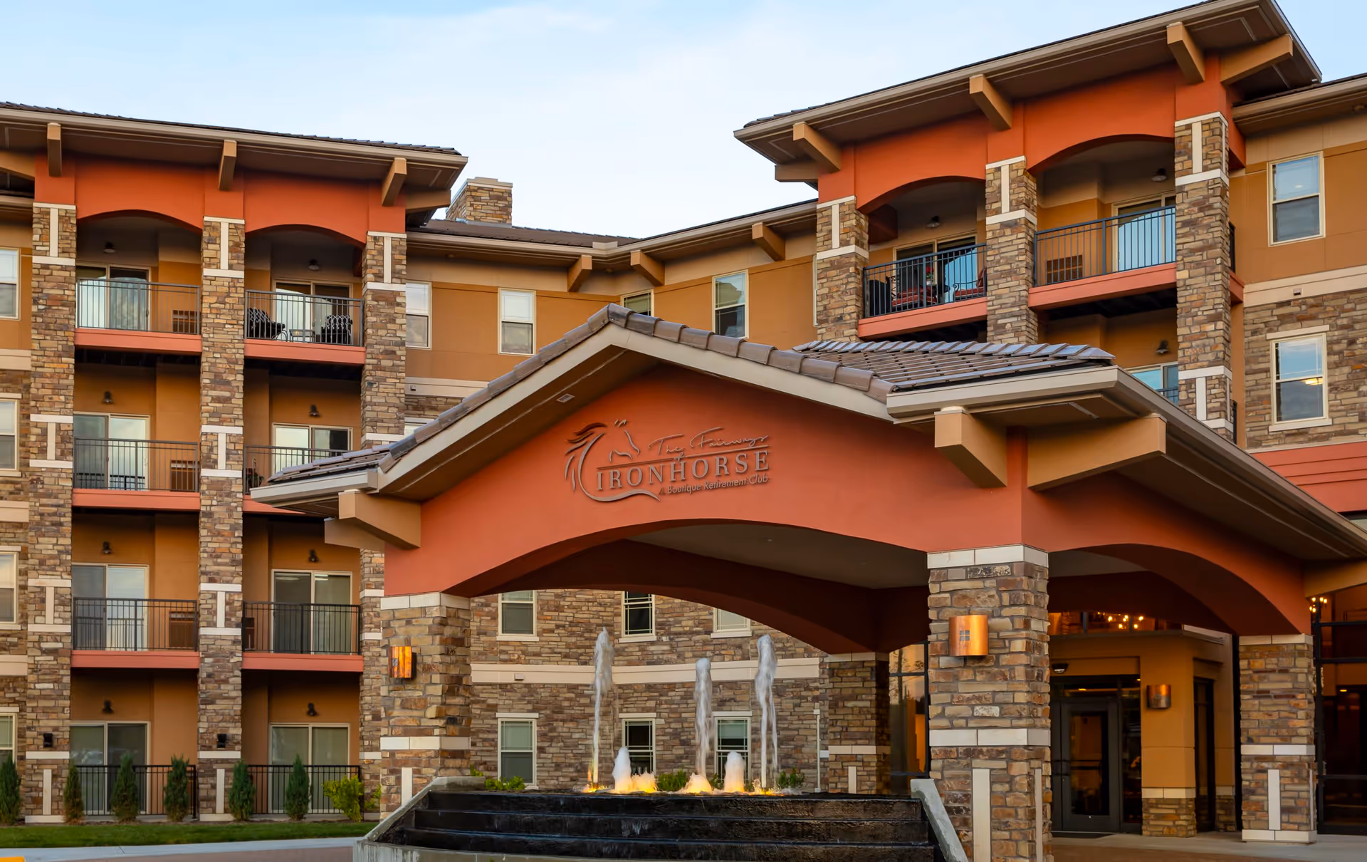 Exterior view of Fairways of Ironhorse Independent Living facility showing a multi-story building with balconies, stone and stucco facade, and a covered entrance with a water fountain in front.