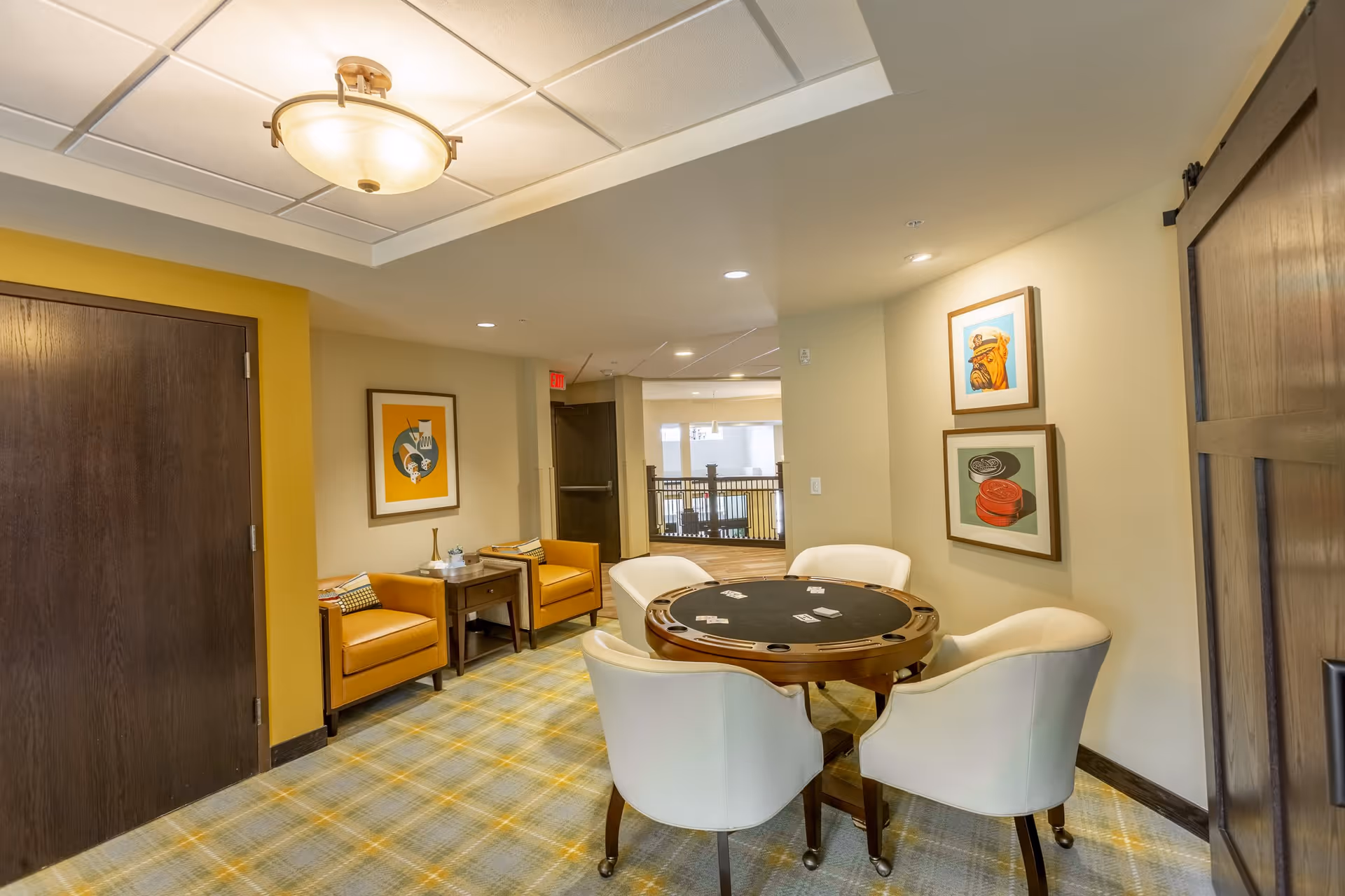 A cozy interior seating area with a round card table surrounded by four white chairs. To the left, there are two orange armchairs separated by a small wooden side table with decorative items. The walls are adorned with framed artwork, and the floor is covered with a patterned carpet. The space is well-lit with ceiling lights and a central ceiling fixture.