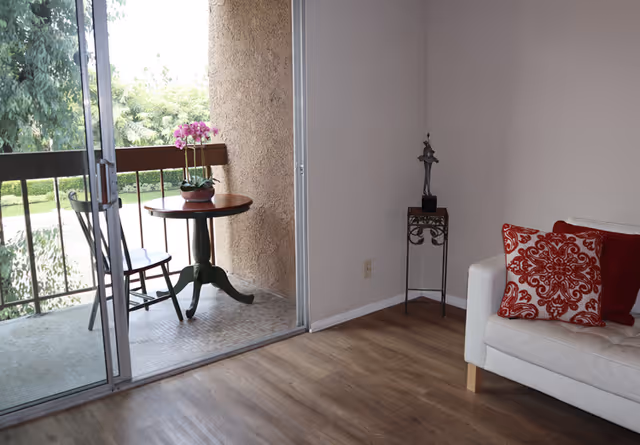 Bright living room corner with a white couch and red patterned pillows opening onto a small balcony with a table, chair, and potted flowers.