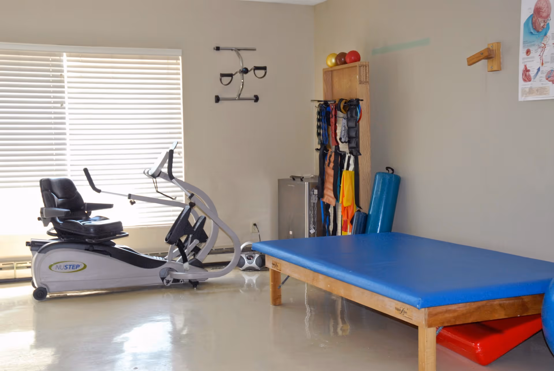 A physical therapy or rehabilitation room with a recumbent exercise bike, a blue padded therapy table, various exercise bands and equipment hanging on a wooden rack, and a medical poster on the wall.