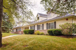 Exterior view of a single-story building with beige siding and brick accents, surrounded by green grass, bushes, and trees under a cloudy sky.
