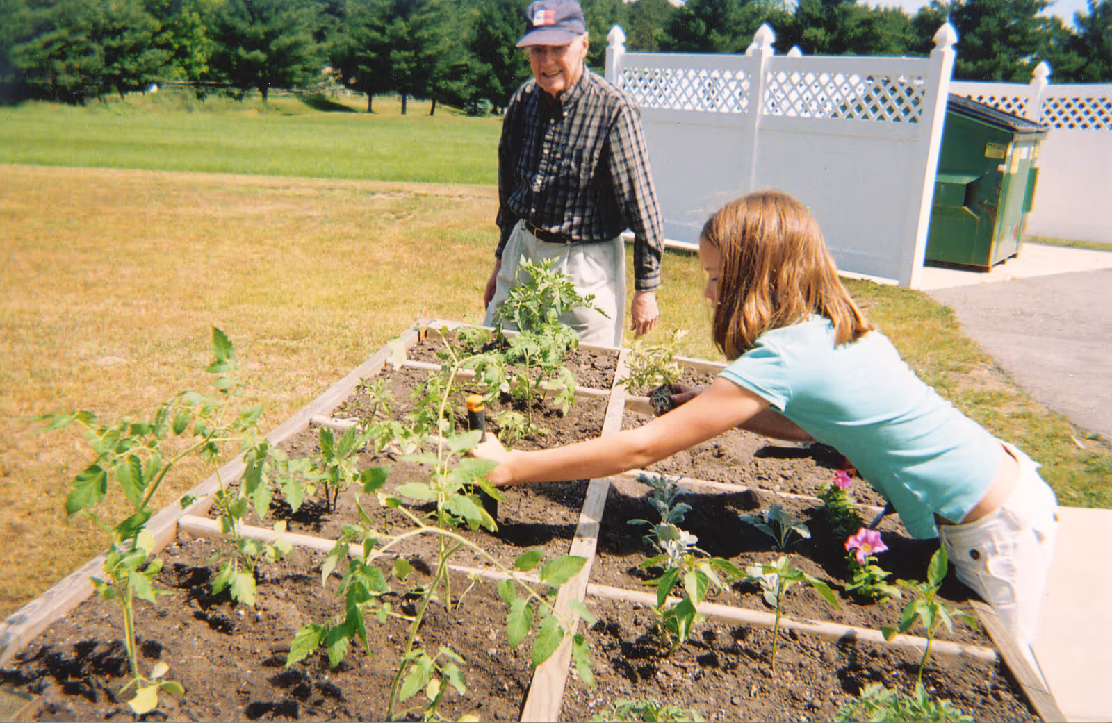 An elderly man and a young girl gardening together in a raised garden bed outdoors. The girl is reaching to tend to the plants while the man watches and smiles. There is a white fence and a green dumpster in the background.