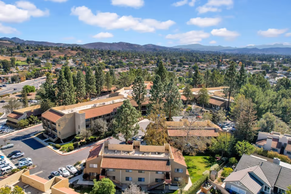 Aerial view of The Reserve At Thousand Oaks senior living facility surrounded by trees and greenery, with multiple buildings featuring terracotta roofs and a parking lot, set against a backdrop of hills and a partly cloudy sky.