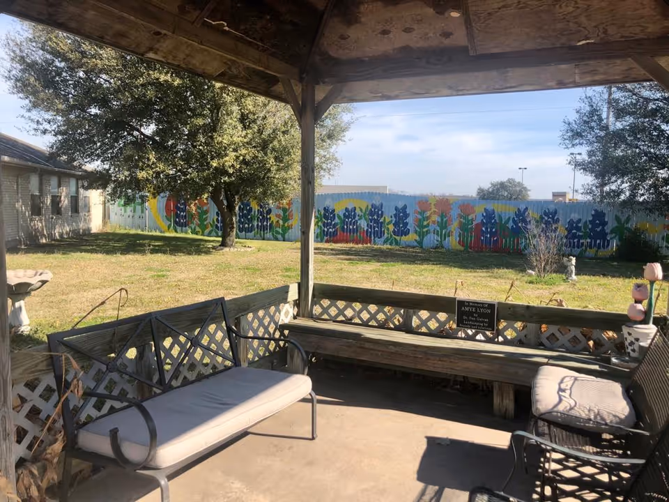 View from a covered outdoor seating area with cushioned benches and chairs, overlooking a grassy yard with trees and a colorful mural painted on a wooden fence in the background.