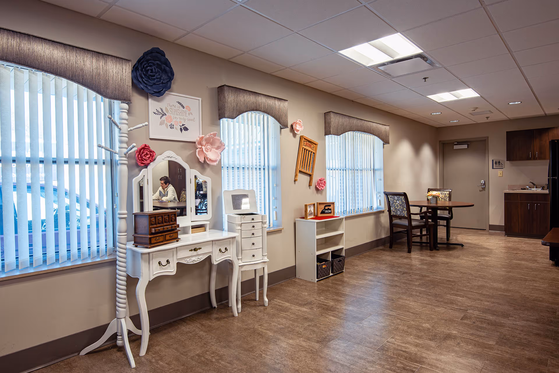 A well-lit interior room with three large windows covered by vertical blinds and valances. Against the wall is a white vintage-style vanity table with a mirror, a small chest of drawers, and a white coat rack adorned with large decorative flowers. There is a small white shelving unit with baskets and framed pictures. In the background, there is a round table with two chairs and a kitchenette area with cabinets and a refrigerator. The floor is wood-patterned, and the ceiling has recessed lighting and fluorescent panels.