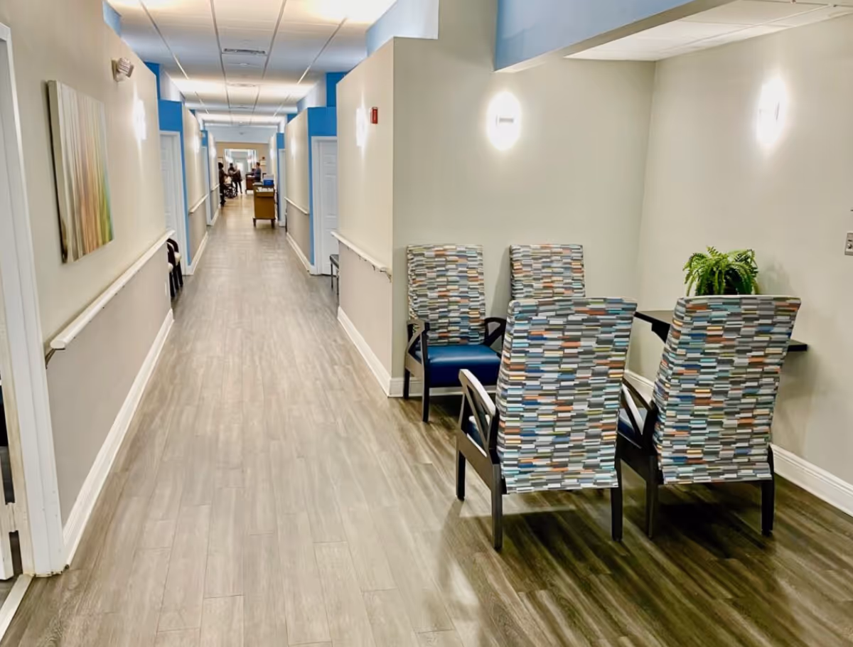 A long, well-lit hallway in an assisted living facility with light-colored walls and wood flooring. On the right side, there is a small seating area with four patterned chairs around a small table with a green plant. The hallway has handrails on both sides and several doors along the walls. In the distance, a few people and a cart are visible.