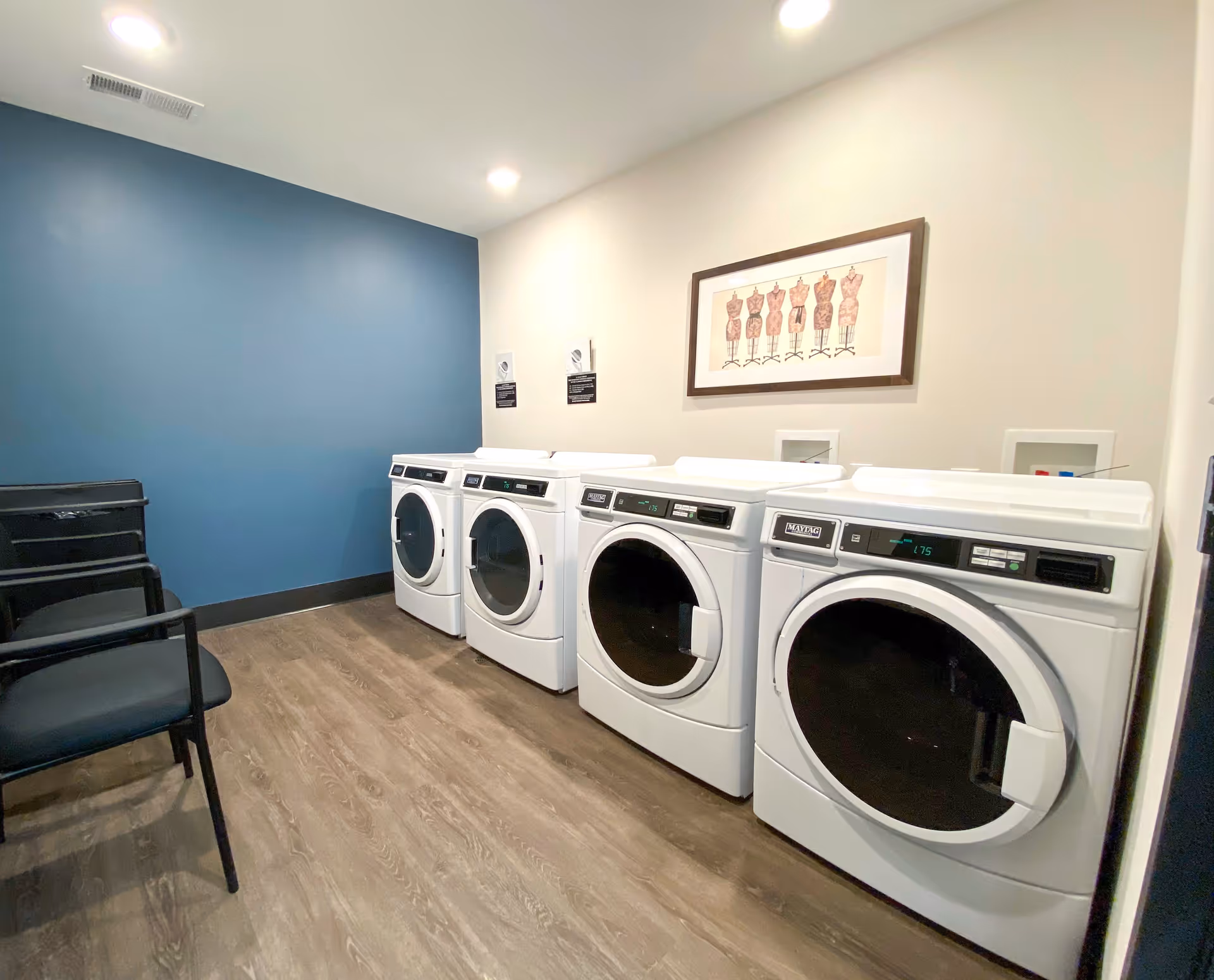 A clean communal laundry room with four front-loading washers/dryers, seating chairs, a blue accent wall, and framed artwork on the wall.