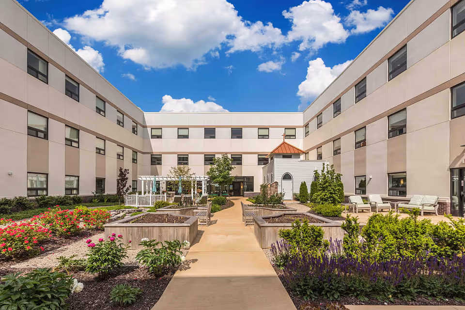 Outdoor courtyard area of The Avalon of Lewis Center facility with raised garden beds, various flowering plants, shrubs, benches, and a small white structure with a copper-colored roof, surrounded by a three-story building under a blue sky with scattered clouds.