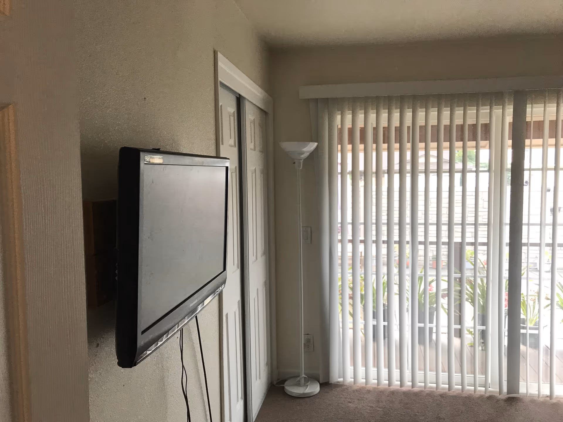 Interior view of a room with a wall-mounted flat screen TV, a white floor lamp, sliding closet doors, and vertical blinds covering a large window or sliding glass door that leads to an outdoor area with plants.