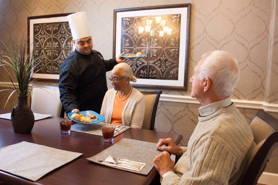 A chef wearing a white hat and black uniform is serving plates of food to two elderly people seated at a dining table in a warmly decorated room with framed artwork and a vase with tall grass on the table.
