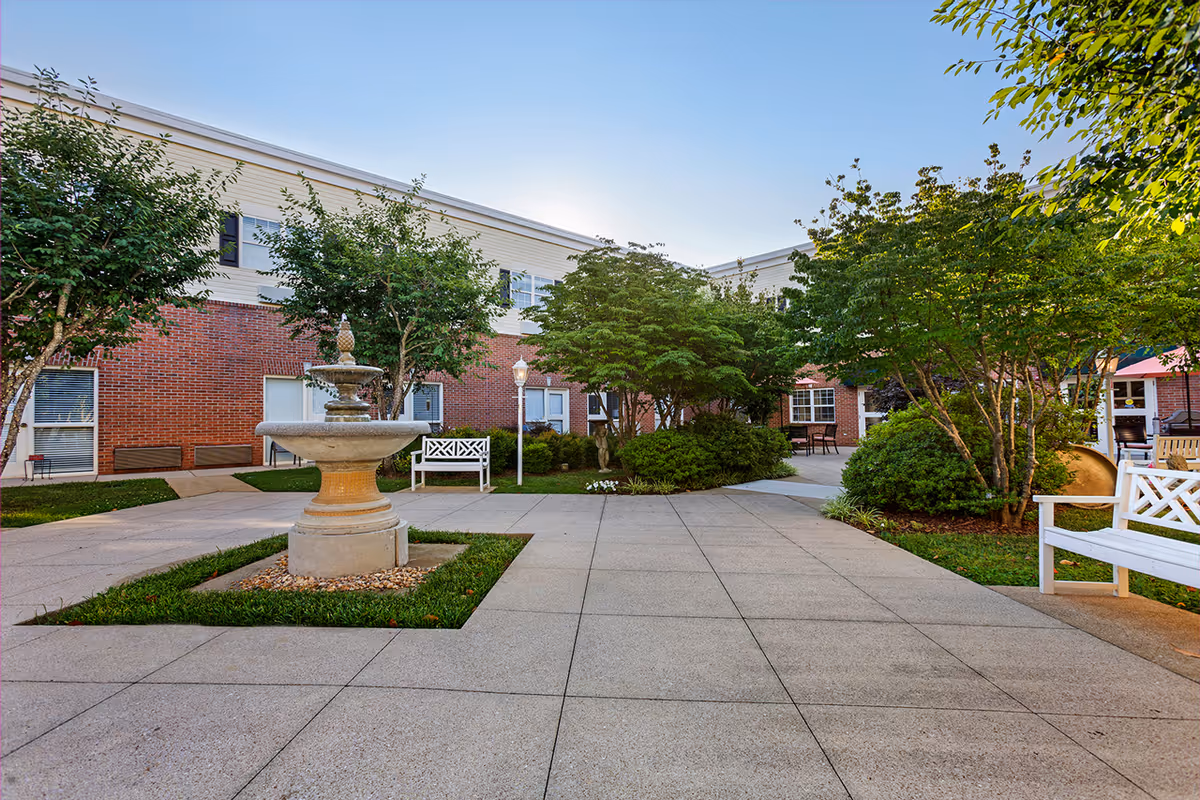 Outdoor courtyard area of a senior living facility with a central stone fountain surrounded by green grass and trees. There are white benches, paved walkways, and a two-story brick and siding building in the background under a clear blue sky.