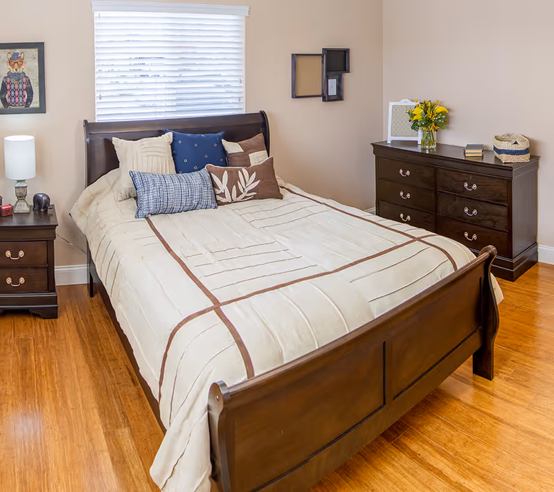 A cozy bedroom with a wooden bed frame and a beige comforter with brown stripes. The bed is adorned with multiple decorative pillows in shades of beige, blue, and brown. To the left of the bed is a wooden nightstand with a lamp, a small elephant figurine, and a red book. On the right side of the room is a wooden dresser with six drawers, topped with a vase of yellow flowers, a small basket, and a couple of books. The walls are painted beige, and there is a window with white blinds behind the bed. A framed picture of a cat wearing glasses and a sweater hangs on the left wall.