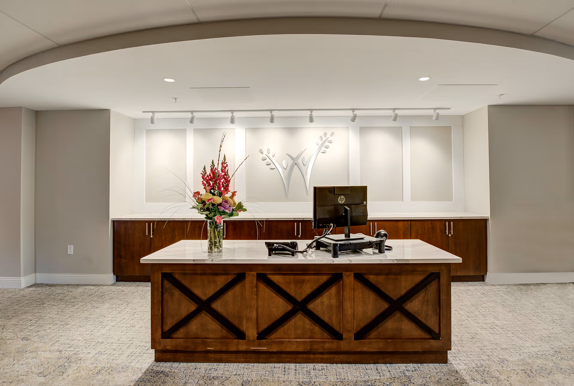 Reception desk area with a wooden counter featuring a vase of flowers and a computer monitor. Behind the desk is a wall with a decorative logo and cabinetry underneath. The space is well-lit with ceiling lights and has a neutral color scheme.