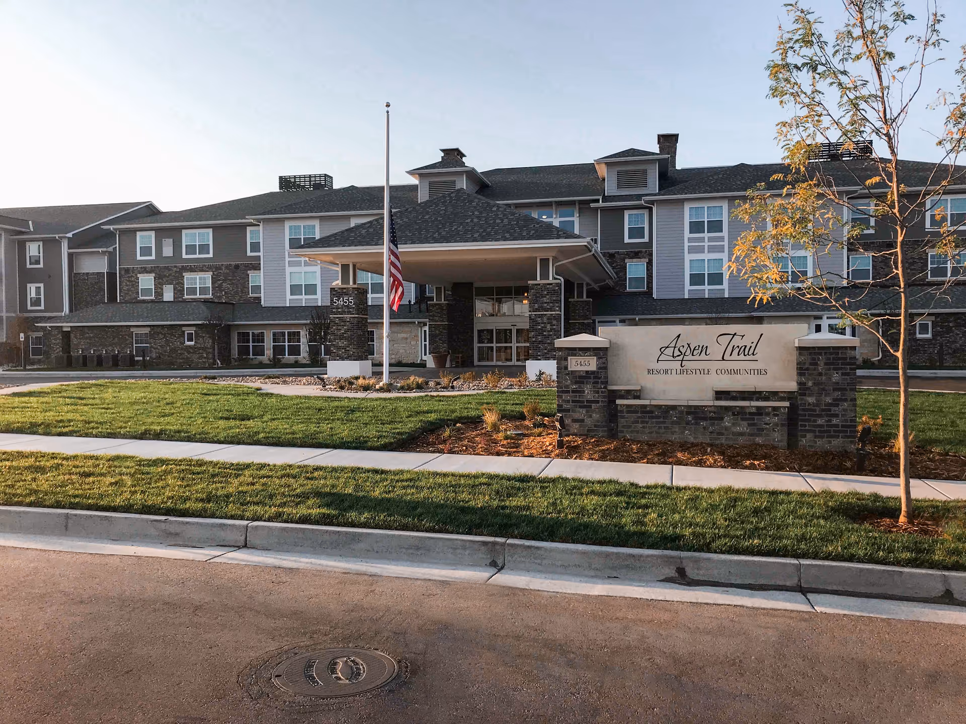 Exterior view of Aspen Trail Retirement Resort building with a well-maintained lawn, a flagpole with an American flag, and a sign displaying the facility's name and address.