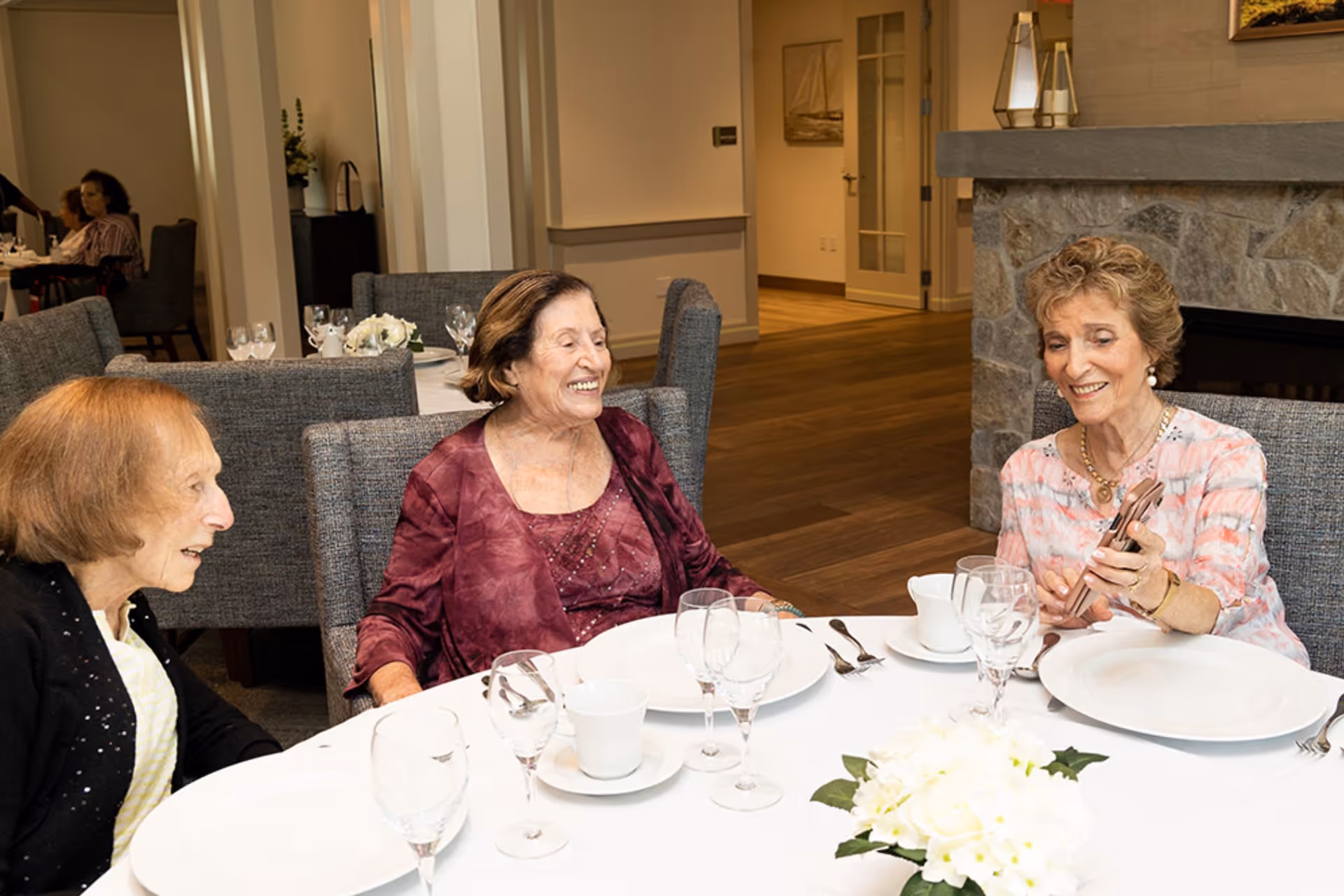 Three elderly women sitting around a dining table in a well-lit room, smiling and engaging with each other. The table is set with white plates, cups, glasses, and silverware, and there is a white floral centerpiece. In the background, other tables and chairs are visible along with a stone fireplace.